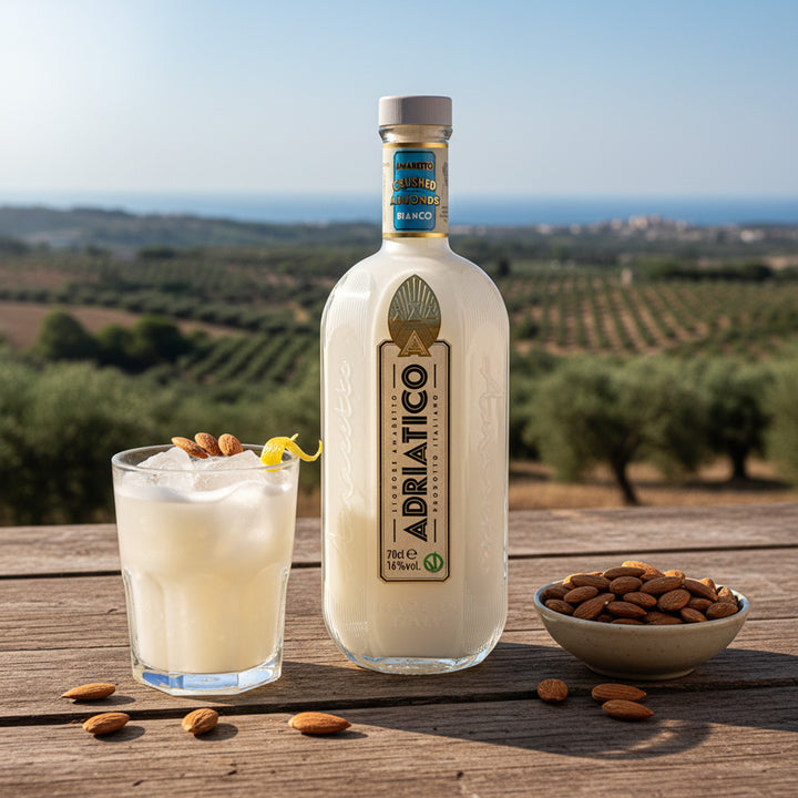 Bottle of Adriatico Amaretto Bianco with a glass of cocktail and almonds on a wooden table with a scenic background.